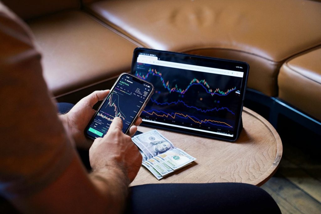 A person using a smartphone and tablet for stock market analysis, with graphs and cash visible.